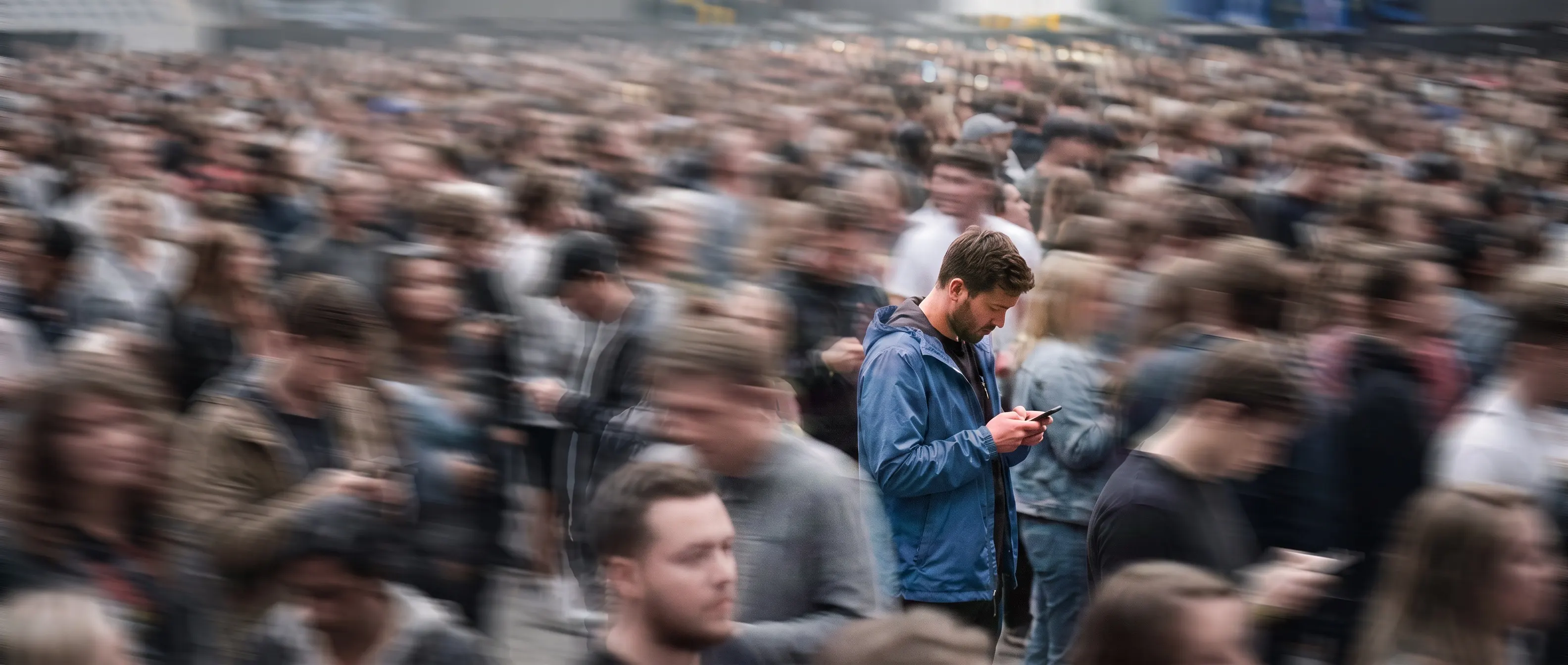 Dichtes Publikum — ein Mann in blauer Jacke steht scharf im Bild und blickt auf sein Smartphone